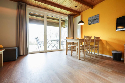 Luminoso salón de lodge con techo de madera, pared amarilla, mesa de comedor y vista a la terraza por puertas de cristal.