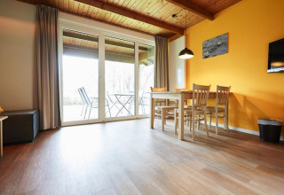 Lyst lodge-opholdsrum med træloft, gule vægge, spisebord, stole og udsigt til terrasse gennem glasdøre.