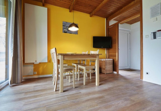 Comedor luminoso en una cabaña con suelo de madera, pared amarilla, mesa, cuatro sillas y TV plana.