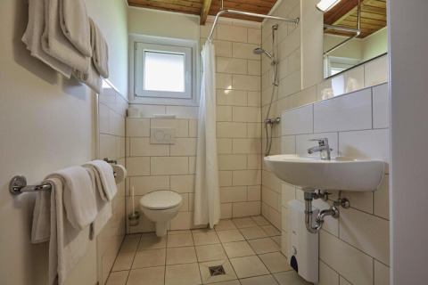 Badeværelse på en lodge med toilet, vask, bruseniche, hvide håndklæder og lyse fliser på væggene.