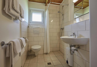 Badeværelse på en lodge med toilet, vask, bruseniche, hvide håndklæder og lyse fliser på væggene.