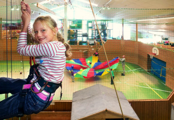 Una niña con arnés se balancea en una sala de juegos cubierta mientras niños juegan con un paracaídas colorido.