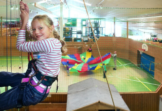 Una ragazza con imbracatura si dondola in un'area giochi coperta, mentre bambini giocano con un paracadute colorato.