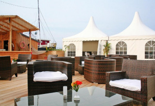 Buitenlounge van een lodge met rotan stoelen, houten terras en witte tenten in de achtergrond.