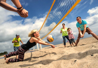 Family enjoying a lively game of beach volleyball by a lodge, having fun on a sandy beach under clouds.