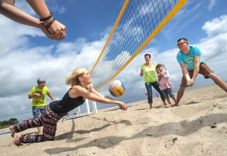Familie spiller beachvolleyball ved en lodge på sandet strand under en skyet himmel med latter og glæde.