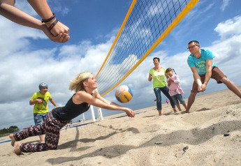 Familie spielt Beachvolleyball an einer Lodge am Sandstrand, fröhliche Stimmung unter bewölktem Himmel.