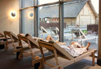Entspannungsbereich im Spa mit Gästen auf Liegen und Blick auf ein Blockhaus durch große Fenster.
