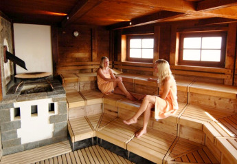 Dos mujeres se relajan y conversan en una sauna revestida de madera con luz natural entrando por las ventanas.