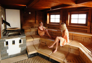 Dos mujeres se relajan y conversan en una sauna revestida de madera con luz natural entrando por las ventanas.