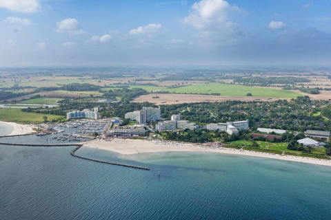 Vista aérea de una zona de cabañas costeras con playa, puerto deportivo, hoteles y campos bajo cielo nublado.