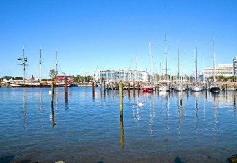 Uitzicht op een jachthaven met zeilboten, historisch schip en lodgebouwen onder een heldere lucht.
