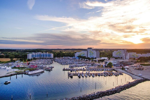 Vista de una marina con barcos y grandes edificios tipo lodge al atardecer, rodeados de naturaleza y campos.