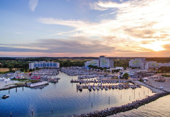 Vue sur une marina avec des bateaux et de grands bâtiments style lodge au coucher du soleil, entourée de nature.