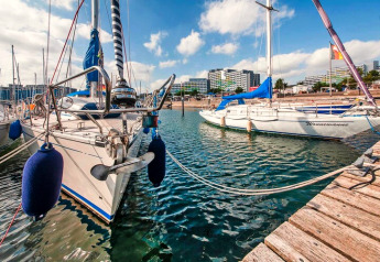 Sailboats docked by a wooden pier at a lodge with modern buildings visible in the background on a sunny day.