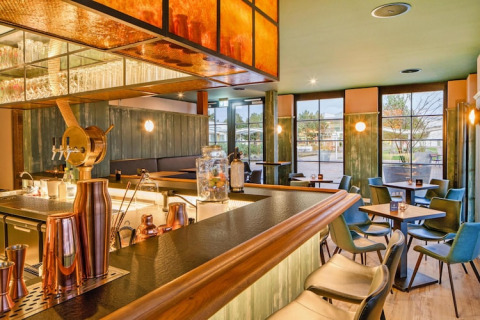 Moderne bar met houten toog en groene stoelen bij Blockhouse HiPr 4 Pers in Duitsland, grote ramen zichtbaar.
