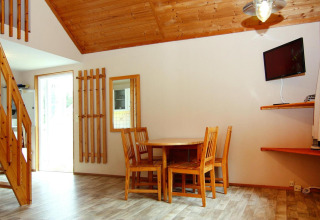 Cozy lodge interior with wooden dining set, staircase, wall-mounted TV, large window, and wood ceiling.