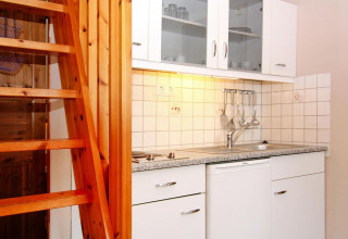 Compact kitchenette with white cabinets, wooden stairs, and utensils at Holiday park Markgrafenheide, Germany.