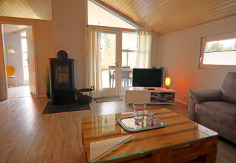 Salon d’une maison de vacances à Hünning, Allemagne, avec poêle, table basse en verre et TV.