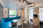 Mobil-home moderne Camping del Mar 6P en Espagne avec canapé bleu, table à manger, TV et large fenêtre.