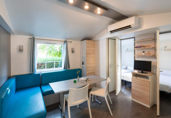 Moderna mobil home Camping del Mar 6P en España con sofá azul, mesa de comedor, TV y ventana luminosa.