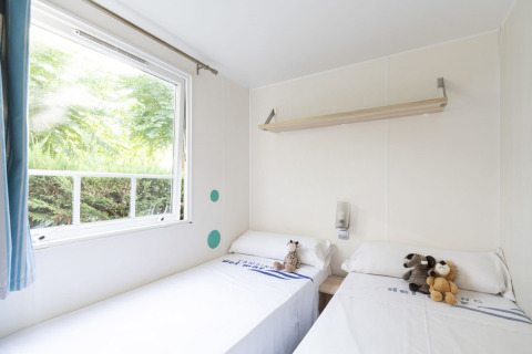 Chambre de mobil-home lumineuse avec deux lits simples, peluches et grande fenêtre donnant sur la verdure.