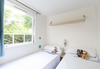 Chambre de mobil-home lumineuse avec deux lits simples, peluches et grande fenêtre donnant sur la verdure.