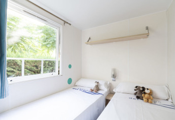 Chambre de mobil-home lumineuse avec deux lits simples, peluches et grande fenêtre donnant sur la verdure.
