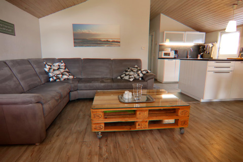 Woonkamer met grijze hoekzetel, pallet salontafel en open keuken in een vakantiewoning in Hünning, Duitsland.