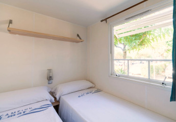 Lichte slaapkamer met twee eenpersoonsbedden, raam en plank in Camping del Mar mobilhome in Spanje.