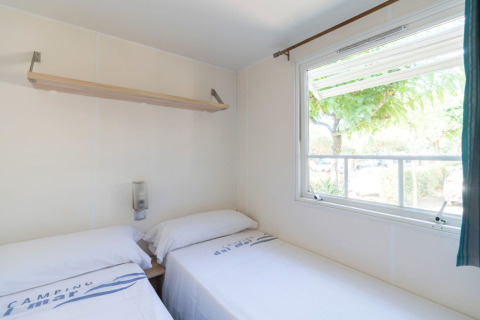 Chambre lumineuse avec deux lits simples, fenêtre et étagère dans le mobil-home Camping del Mar en Espagne.