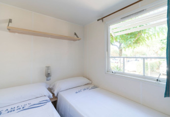 Chambre lumineuse avec deux lits simples, fenêtre et étagère dans le mobil-home Camping del Mar en Espagne.
