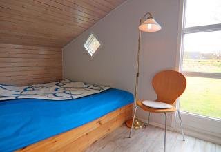 Camera accogliente con soffitto inclinato in legno, letto blu, sedia e lampada da terra in una casa vacanze a Hünning, Germania.