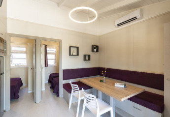Comedor moderno con mesa de madera, banco esquinero púrpura y sillas blancas en Camping del Mar BUNGALOW PREMIUM, España.