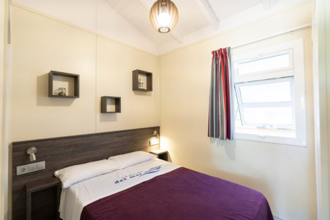 Helles Schlafzimmer mit Doppelbett, Fenster, lila Decke und moderner Dekoration im Camping del Mar BUNGALOW PREMIUM in Spanien.