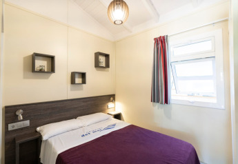 Dormitorio luminoso con cama doble, ventana, manta morada y decoración moderna en Camping del Mar BUNGALOW PREMIUM en España.