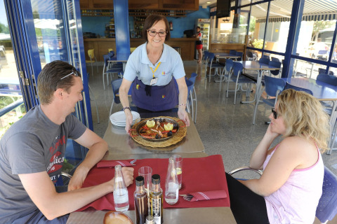 Eine Kellnerin bringt köstliches Essen zu zwei Gästen auf der Terrasse vom Camping del Mar BUNGALOW PREMIUM in Spanien.