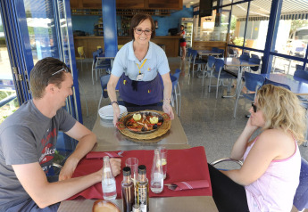 Una camarera sirve un plato de mariscos a dos personas sentadas al aire libre en Camping del Mar BUNGALOW PREMIUM, España.