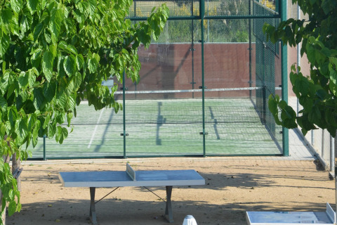 Udendørs bordtennisbord under træer og indhegnet padelbane i baggrunden ved en mobilhome-plads.
