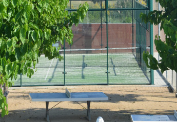 Mesa de ping-pong al aire libre bajo árboles y pista de pádel vallada al fondo en un mobil home.
