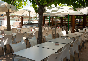 Zona exterior de comedor en glamping, con sillas blancas, mesas, sombrillas y árboles con follaje.