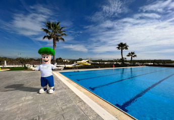Mascot posing by outdoor swimming pool at Els Pins- Mobile-Home Plus 3 in Spain on a sunny day with palm trees.