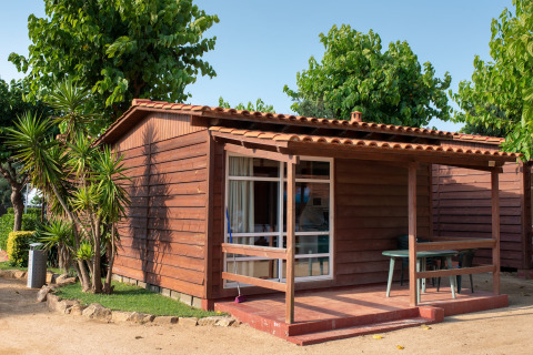 Wooden bungalow with veranda at Els Pins- Bungalow Comfort, surrounded by lush greenery and trees.