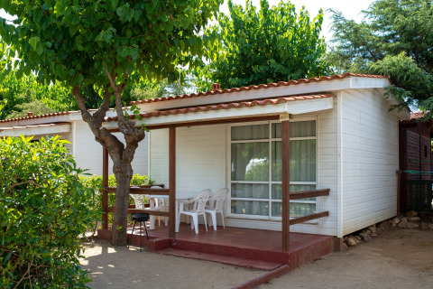 Outdoor view of Els Pins- Bungalow Comfort lodge in Spain, showing a porch, chairs and lush trees.