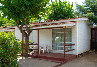 Outdoor view of Els Pins- Bungalow Comfort lodge in Spain, showing a porch, chairs and lush trees.