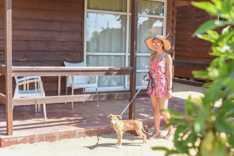 Woman in sunhat with dog on leash standing outside wooden bungalow at Els Pins- Bungalow Comfort, Spain.