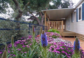 Stacaravan bij Camping Rocagrossa in Spanje met bloemenperk en houten veranda naast een hek.
