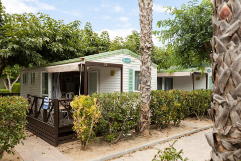 Mobilhjem Camping Cambrils Playa BUNGALOW OHARA med terrasse og omgivet af grønne buske og palmetræer.