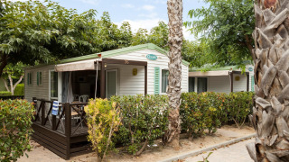 Mobilhjem Camping Cambrils Playa BUNGALOW OHARA med terrasse og omgivet af grønne buske og palmetræer.