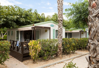 Stacaravan Camping Cambrils Playa BUNGALOW OHARA met veranda, omgeven door palmbomen en groene struiken.